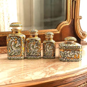 Puede incluir: Un conjunto de cuatro recipientes de cerámica con base cuadrada o hexagonal y tapa a juego. Cada pieza está decorada con un patrón floral repetitivo en tonos verdes y blancos, acentuado con bordes dorados en los extremos y las tapas.