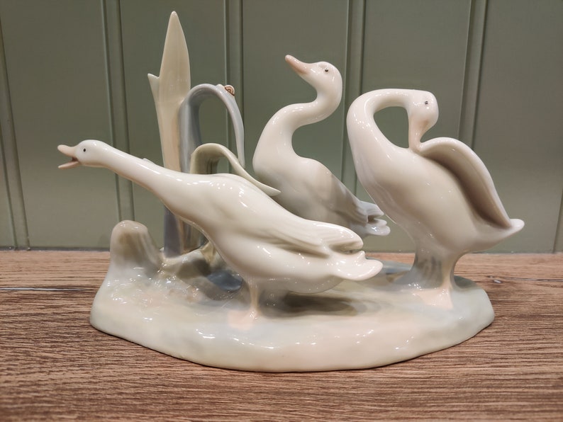 May include: A porcelain figurine of three white geese standing on a base with a green plant. The geese are in a variety of poses, with one looking up, one looking down, and one looking to the side.