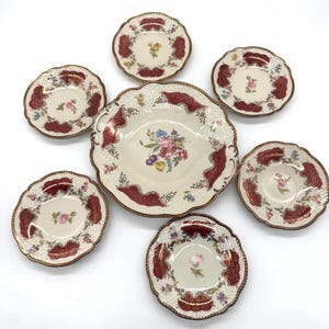 May include: A set of antique porcelain dishes. The set includes a large serving bowl and six smaller plates. The dishes feature a cream-colored background with burgundy and gold trim, and floral designs.