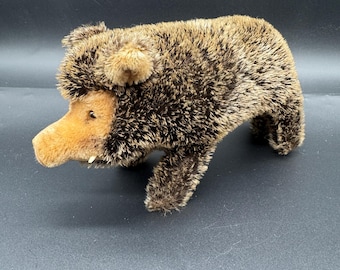 May include: A plush toy boar with brown and tan fur. The boar has a tan snout, small tusks, and small ears. The toy is standing on all four legs.