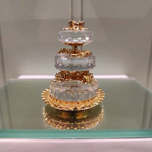 以下が含まれることがあります： 金色のアクセントが施された3段のクリスタルケーキの置物。ケーキは複雑なディテールで飾られ、頂上には王冠が置かれています。
