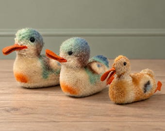 May include: Three plush ducklings with soft, fuzzy textures. The two larger ducks feature cream, blue, and green plumage with bright orange beaks and chest patches. The smallest duck is primarily golden-brown with a matching orange beak.