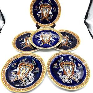 May include: A set of six decorative ceramic plates. The plates feature a dark blue background with a detailed central design in gold, white, and orange. The plates have a gold border with a repeating pattern.
