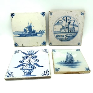 May include: Four square ceramic tiles featuring blue and white hand-painted designs. One tile shows a windmill, another a building in a circle, one a floral vase, and the last a sailboat on water. Each tile has small blue corner motifs.