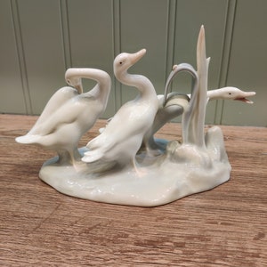 May include: A porcelain figurine of three white ducks standing on a base with a green plant. The ducks are in a playful pose, with one duck looking back at the viewer.