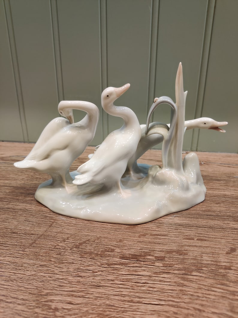 May include: A porcelain figurine of three white ducks standing on a base with a green plant. The ducks are in a playful pose, with one duck looking back at the viewer.