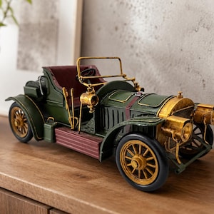 May include: A vintage-style metal model car in dark green with gold-toned wheels, headlights, and radiator grille. The vehicle features a maroon interior, a thin gold windscreen frame, and black tyres, capturing the aesthetic of an early automobile.