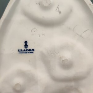 May include: The bottom of a white porcelain figurine with a blue and white Lladro logo that reads "Lladro Hand Made in Spain". The figurine is marked with the number "416" and the number "4" is written in pencil.