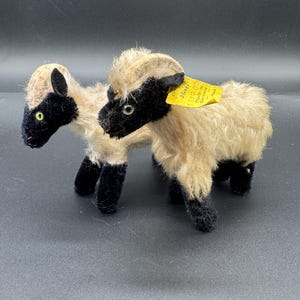 Puede incluir: Dos ovejas de juguete vintage con cuerpos de lana beige y negra. Las ovejas tienen caras, orejas y patas negras, con etiquetas de precio amarillas. Las ovejas están sobre una superficie oscura.