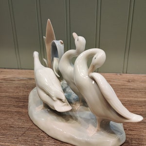 May include: A porcelain figurine of three white geese standing on a base. The geese are arranged in a playful pose, with one goose looking back at the other two.