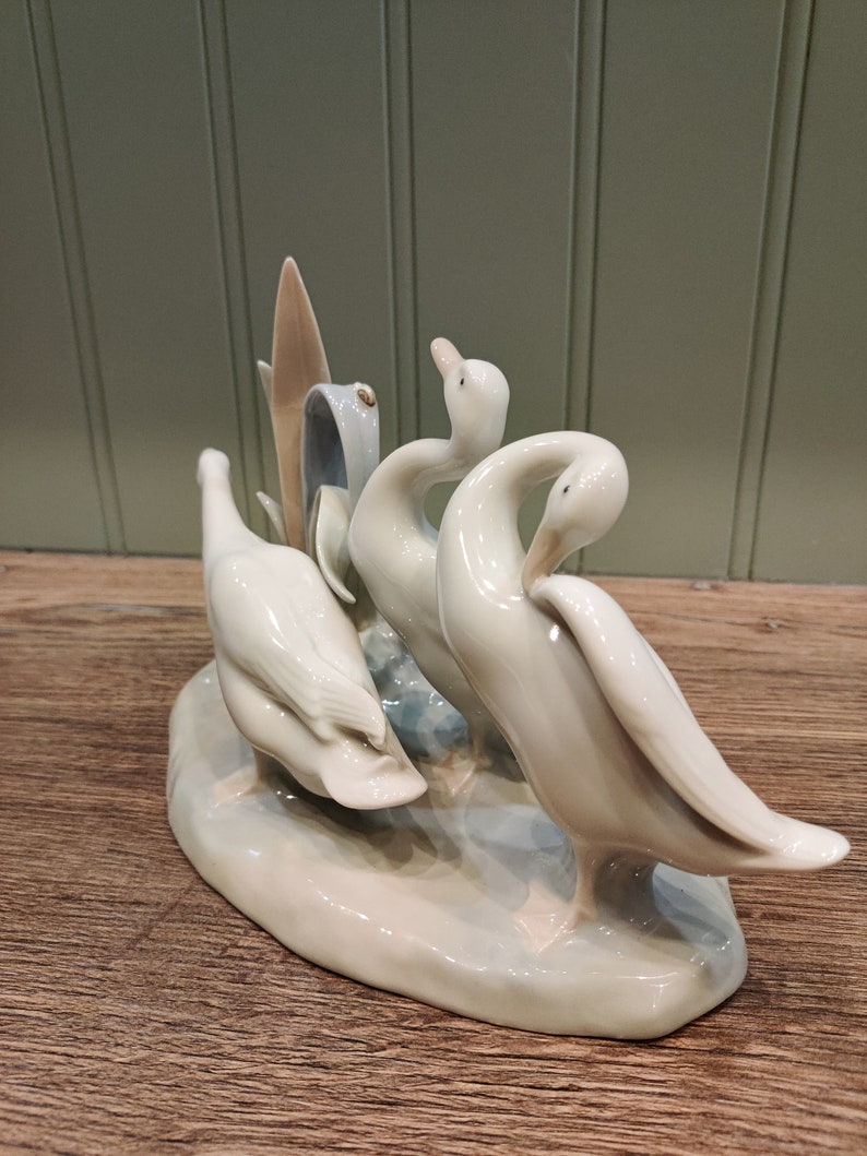 May include: A porcelain figurine of three white geese standing on a base. The geese are arranged in a playful pose, with one goose looking back at the other two.