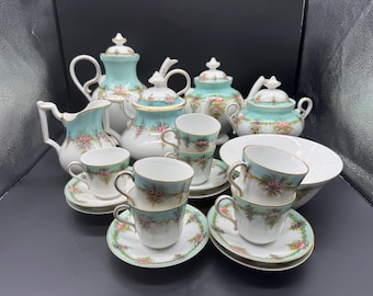 May include: A vintage tea set featuring teacups, saucers, teapots, a creamer, and a serving bowl. The set has a white base with a teal border and floral accents. The teacups and saucers are stacked, and the teapots have lids.