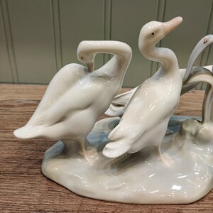 May include: A porcelain figurine of two white geese standing on a base. The geese are in a playful pose, with one goose's head tucked under its wing.