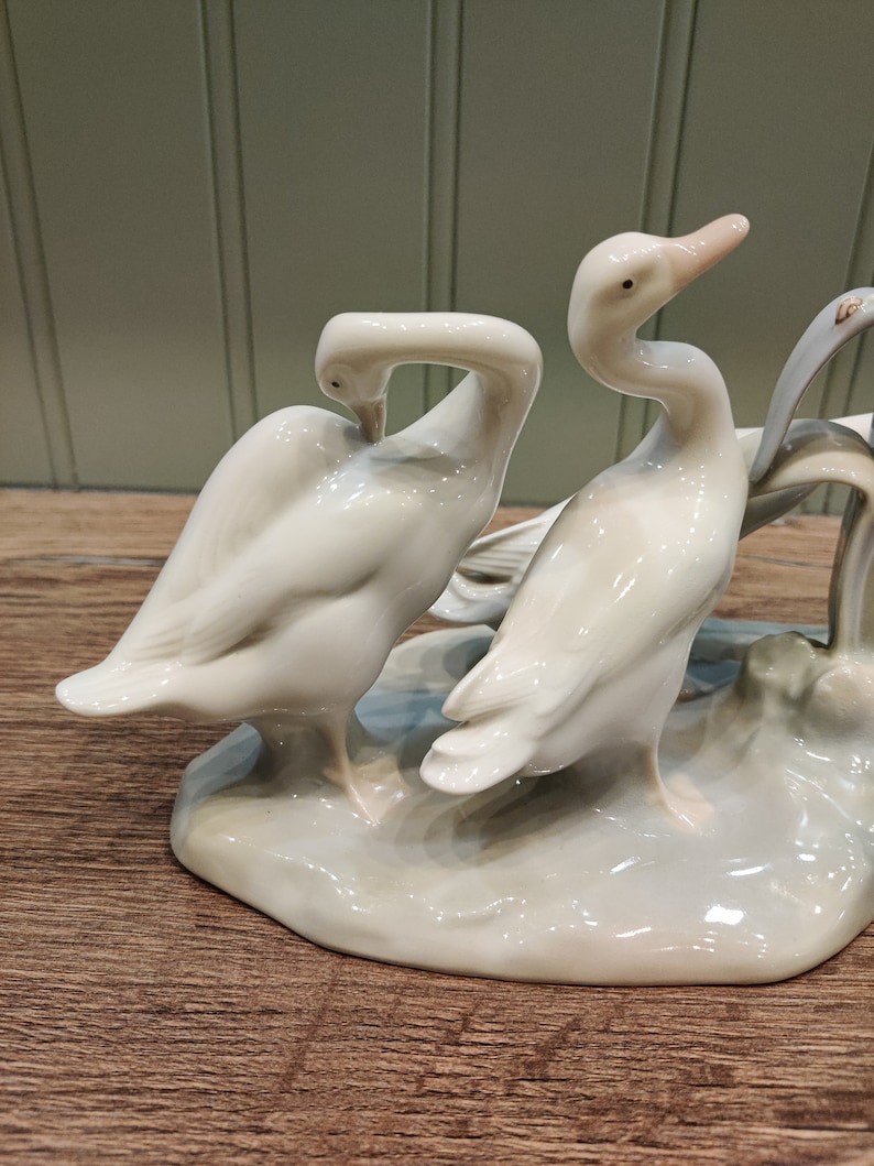 May include: A porcelain figurine of two white geese standing on a base. The geese are in a playful pose, with one goose's head tucked under its wing.