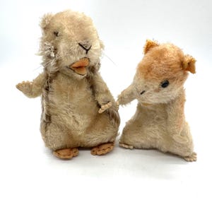 May include: Two vintage stuffed animal hamsters. The larger hamster is light brown with a darker brown patch on its back. The smaller hamster is light brown with orange ears. Both have black eyes and are made of a soft, plush material.