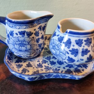 May include: A blue and white porcelain creamer and sugar bowl set with a matching tray. The set features a floral design with blue flowers and leaves on a white background.
