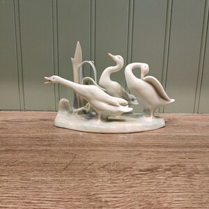 May include: A porcelain figurine of three white geese standing on a base with a green and blue background. The geese are in a natural pose, with their heads turned and their wings spread.