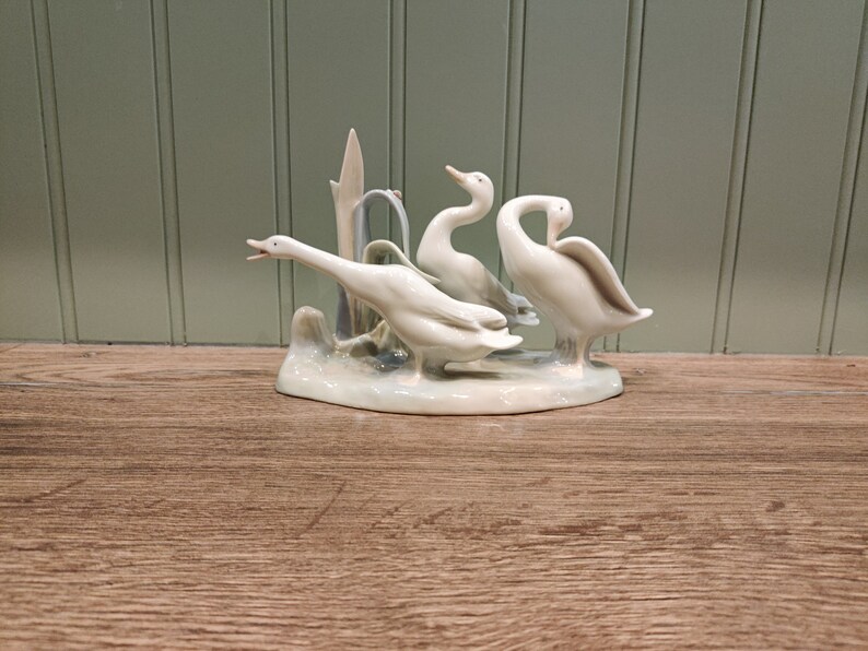 May include: A porcelain figurine of three white geese standing on a base with a green and blue background. The geese are in a natural pose, with their heads turned and their wings spread.