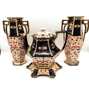 Könnte beinhalten: Ein Vintage-Teeservice mit einer Teekanne und zwei Vasen. Das Set hat eine cremefarbene Basis mit roten und schwarzen Akzenten, goldfarbenen Verzierungen und einem floralen Muster. Die Teekanne steht auf einem dekorativen Sockel.