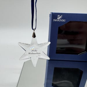 Puede incluir: Un adorno navideño de cristal transparente en forma de estrella con el texto "Frohe Weihnachten" suspendido por una cinta azul marino. Una caja azul marino con el logotipo de Swarovski está en el fondo. Decoración festiva.