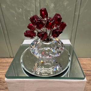 Può includere: Un vaso di cristallo con un bouquet di rose rosse. Il vaso è trasparente e ha una base rotonda. Le rose sono di cristallo e hanno un colore rosso. Il vaso è appoggiato su una base a specchio. La base ha il testo "Swarovski" e "2002" su di essa. La base è appoggiata su una scatola bianca con il testo "VDF Antiques & Collectibles" su di essa.