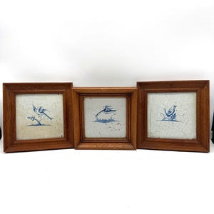 May include: Three square ceramic tiles framed in medium-toned wood. Each tile features a white background with a visible crackle glaze finish and a blue hand-painted illustration of a bird perched on a branch or foliage in a distinct pose.