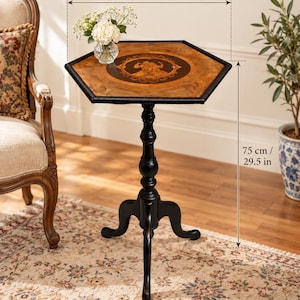 May include: A hexagonal wooden side table with a dark brown finish, featuring a central inlay design on the tabletop. The table stands on a single pedestal base with three curved legs. It measures 46 x 39 cm in width and 75 cm in total height.