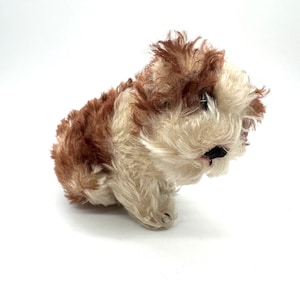 Puede incluir: Un perro de peluche vintage con pelaje marrón y crema. El animal de peluche está en posición sentada, con la cabeza inclinada hacia abajo. El juguete tiene una nariz negra y ojos pequeños y oscuros. El pelaje del perro es esponjoso y texturizado.