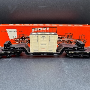 May include: A model railway flat wagon carrying a beige container marked "BAHIA" with an umbrella symbol. The black wagon has detailed features, and a red box with German text is in the background.