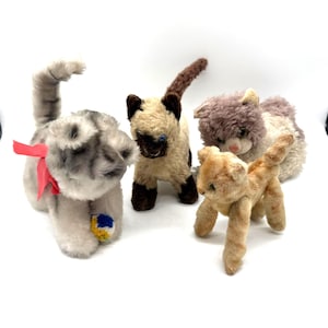 May include: Four vintage stuffed cat toys in various colors. One is gray with a red bow, another is a Siamese cat, and the others are tan and lavender. Each cat has a unique pose and expression, showcasing their individual charm.