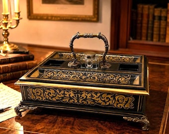 May include: A rectangular black desk organizer featuring intricate gold scrollwork patterns, a central metal handle, and a clear glass inkwell. The piece is supported by four ornate metal feet and includes multiple compartments for stationery storage.