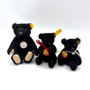 Puede incluir: Tres osos de peluche negros con patas color canela y pequeñas etiquetas amarillas. Los osos tienen ojos negros de botón y están sentados. Un oso tiene una cinta roja y otro una cinta blanca. Un oso tiene una etiqueta circular que dice "Teddybar 1912".