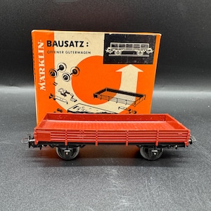 Op de afbeelding: Een rode modeltrein platte wagen met zwarte wielen en accenten staat voor zijn oranje en witte doos. Op de doos staat "BAUSATZ: OFFENER GUTERWAGEN" en een diagram van de wagon.