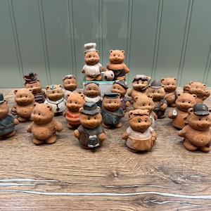 Può includere: Una collezione di 18 piccole figurine di orso in ceramica marrone, ciascuna con un abito e un cappello diversi. Gli orsi sono disposti a semicerchio su una superficie di legno.