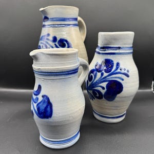 Può includere: Tre brocche in ceramica con disegni floreali blu. Le brocche sono di varie dimensioni, con un colore di base beige chiaro e bianco. Ogni brocca ha un manico e un beccuccio, con fasce e motivi floreali blu.