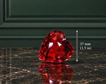 Puede incluir: Una piedra preciosa roja con forma de corazón y corte brillante facetado. Un indicador de medida vertical muestra que el objeto tiene 37 mm de altura. La piedra muestra tonos carmesí profundos y facetas geométricas que reflejan la luz.