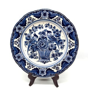 Pode incluir: Um prato de cerâmica decorativo com um design floral azul e branco. O prato apresenta uma imagem central de dois pássaros empoleirados acima de um vaso de flores. A borda é adornada com um padrão floral repetitivo. O prato é exibido em um suporte de madeira marrom.