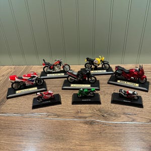 Könnte beinhalten: Eine Sammlung von sieben Miniatur-Motorradmodellen auf schwarzen Präsentationsständern. Die Modelle sind in verschiedenen Farben, darunter Rot, Schwarz, Grün, Gelb und Weiß. Die Modelle zeigen verschiedene Marken, darunter Honda, Kawasaki und BMW.