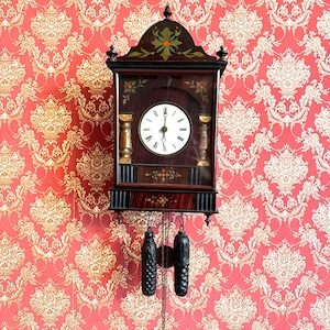 Può includere: Un orologio da parete in legno scuro con quadrante bianco e lancette nere. L'orologio ha dettagli dorati decorativi e due pesi neri appesi sotto. L'orologio è montato su una carta da parati rossa e oro.