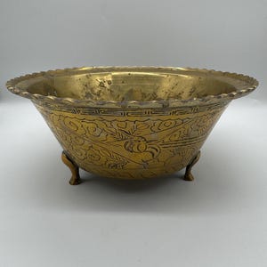 May include: A vintage brass bowl with a detailed floral pattern and a scalloped rim. The bowl rests on three small feet. The brass has a warm, aged patina, adding to its antique charm. This decorative bowl is suitable for display or functional use.