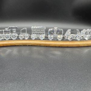 May include: A clear crystal train set with a locomotive, passenger cars, and caboose. The train is mounted on a curved wooden base. The train cars are connected with small metal chains. The background is dark.