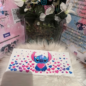 May include: A blue Stitch character holding a pink heart surrounded by pink and purple hearts on a white background. The text "Morena's Creations And More" is visible in the background.