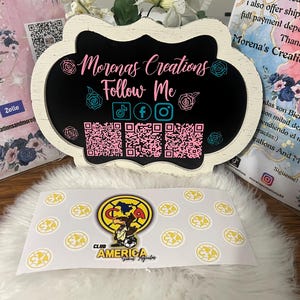 May include: A white sign with a black chalkboard insert that says "Morenas Creations Follow Me" with social media icons and QR codes. The sign is sitting on a white fluffy surface with yellow stickers that say "Club America" with a cartoon eagle logo.