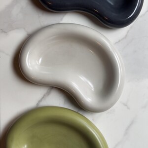 May include: Three ceramic bowls in different colors: green, white, and gray. The bowls are shaped like a kidney bean and have a smooth, glossy finish.