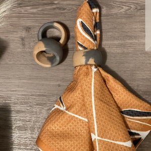 K&ouml;nnte beinhalten: Zwei graue und braune Serviettenringe mit geometrischem Muster auf einer braunen und wei&szlig;en gemusterten Serviette.