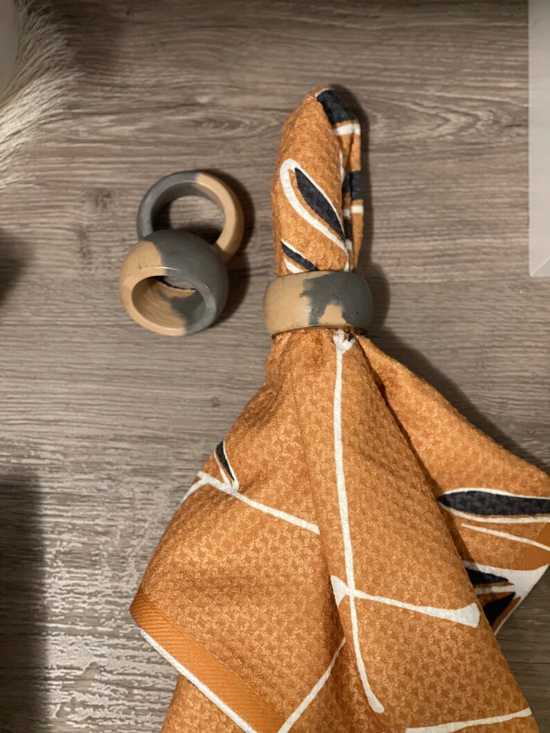 K&ouml;nnte beinhalten: Zwei graue und braune Serviettenringe mit geometrischem Muster auf einer braunen und wei&szlig;en gemusterten Serviette.