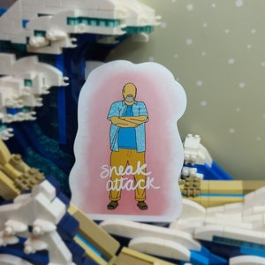 May include: A sticker featuring a cartoon man with arms crossed, wearing a blue shirt and yellow pants. The sticker has the text "sneak attack" in white script. The background is a pink cloud.