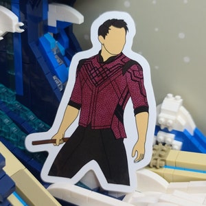 May include: A sticker featuring a character in a maroon and black outfit, holding a brown staff. The character has a tan face and black hair. The sticker is set against a background of a blue and white wave made of building blocks.
