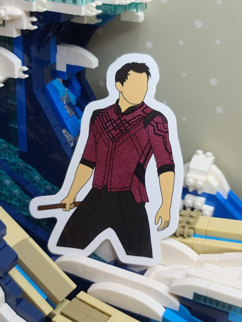 May include: A sticker featuring a character in a maroon and black outfit, holding a brown staff. The character has a tan face and black hair. The sticker is set against a background of a blue and white wave made of building blocks.