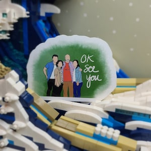 May include: A sticker featuring a family portrait with the text "OK See you" in a handwritten style. The sticker is set against a backdrop of a Lego wave, with shades of blue, white, and tan.